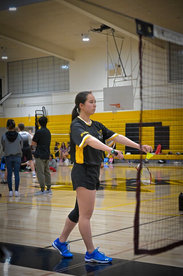 A person in sports attire is standing on a badminton court, preparing to play The gymnasium has a yellow and black color scheme, with other people and equipment visible in the background